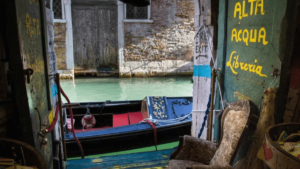 O interior da entrada da "Alta Acqua Libreria" em Veneza, mostrando a porta aberta com o nome da livraria pintado em amarelo e uma gôndola de Veneza atracada bem ao lado, visível através da abertura.