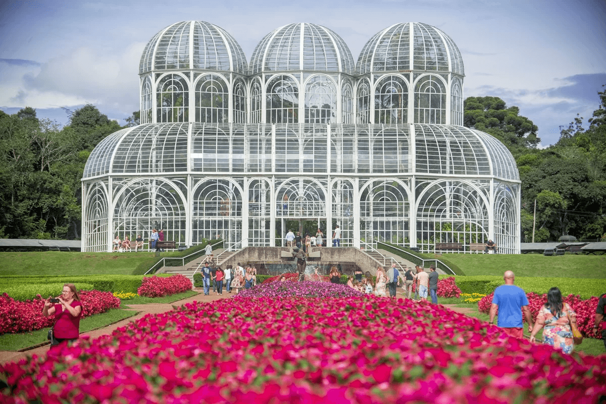 Uma imagem da icônica estufa do Jardim Botânico de Curitiba. A estrutura de metal e vidro é cercada por jardins vibrantes de flores pink. Turistas podem ser vistos passeando e apreciando as plantas ao redor da estufa, que é uma das principais atrações turísticas da cidade.