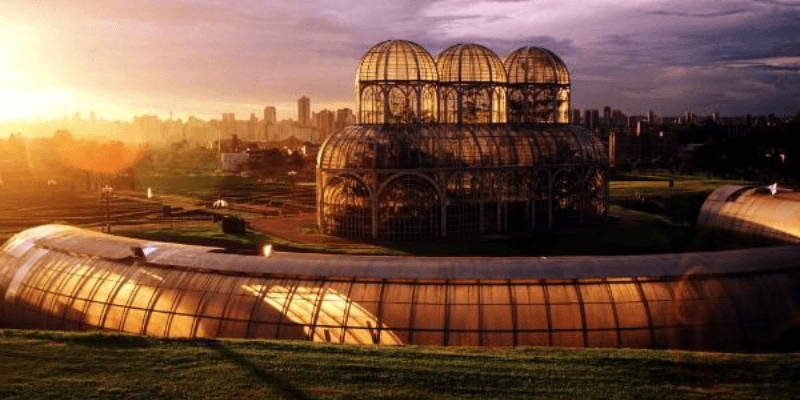 O sol dourado do entardecer ilumina a icônica estrutura de metal e vidro do Jardim Botânico de Curitiba, destacando a simetria e beleza do espaço ao pôr do sol.