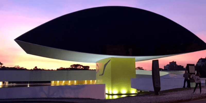 Vista do Museu Oscar Niemeyer ao pôr do sol em Curitiba, com o céu colorido em tons de rosa e azul, e iluminação destacando sua forma única, semelhante a um olho.