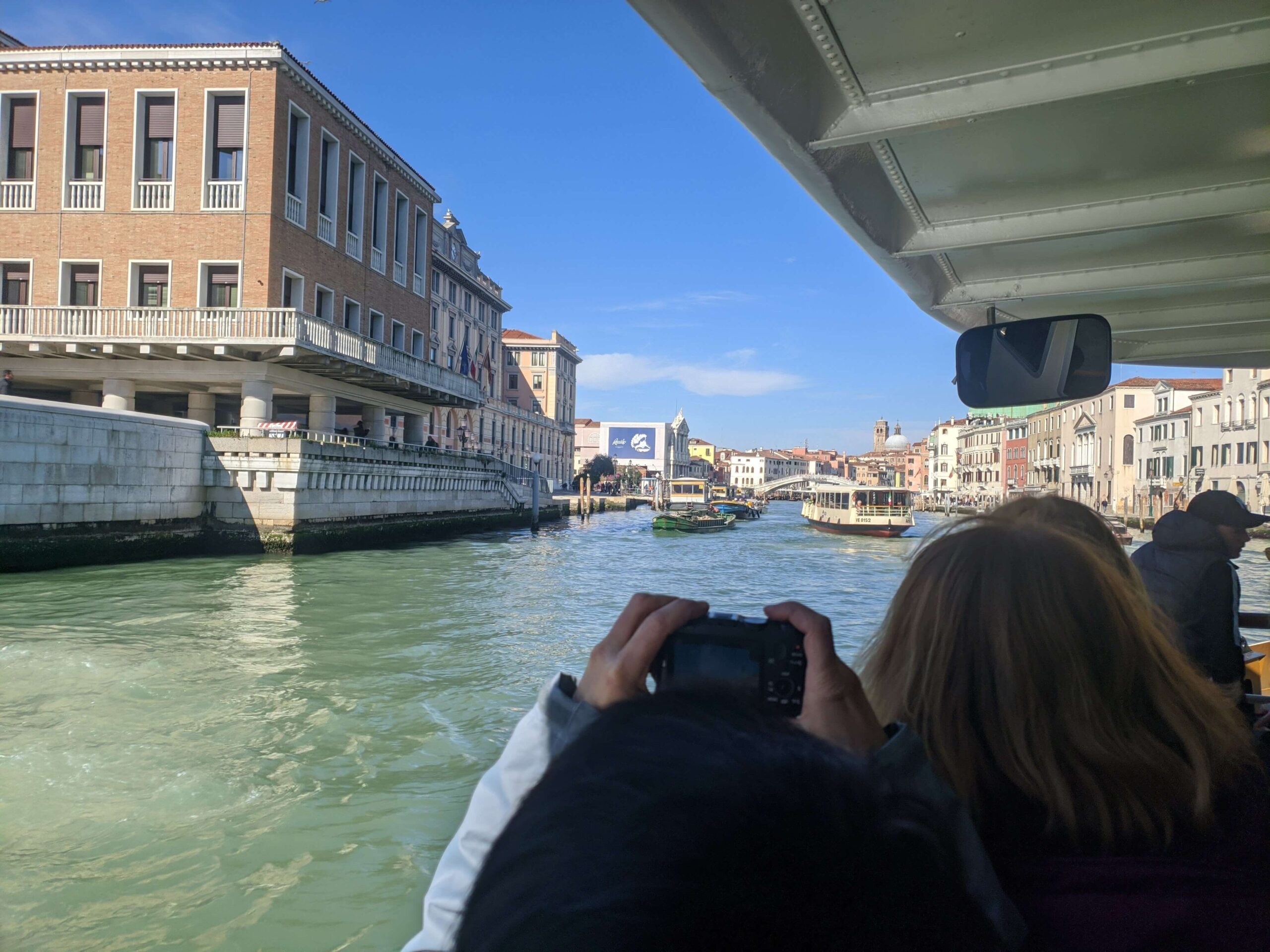 Vista de um passeio de vaporetto em Veneza, com passageiros à frente fotografando a vista do Grande Canal e sua arquitetura fascinante e barcos passando.