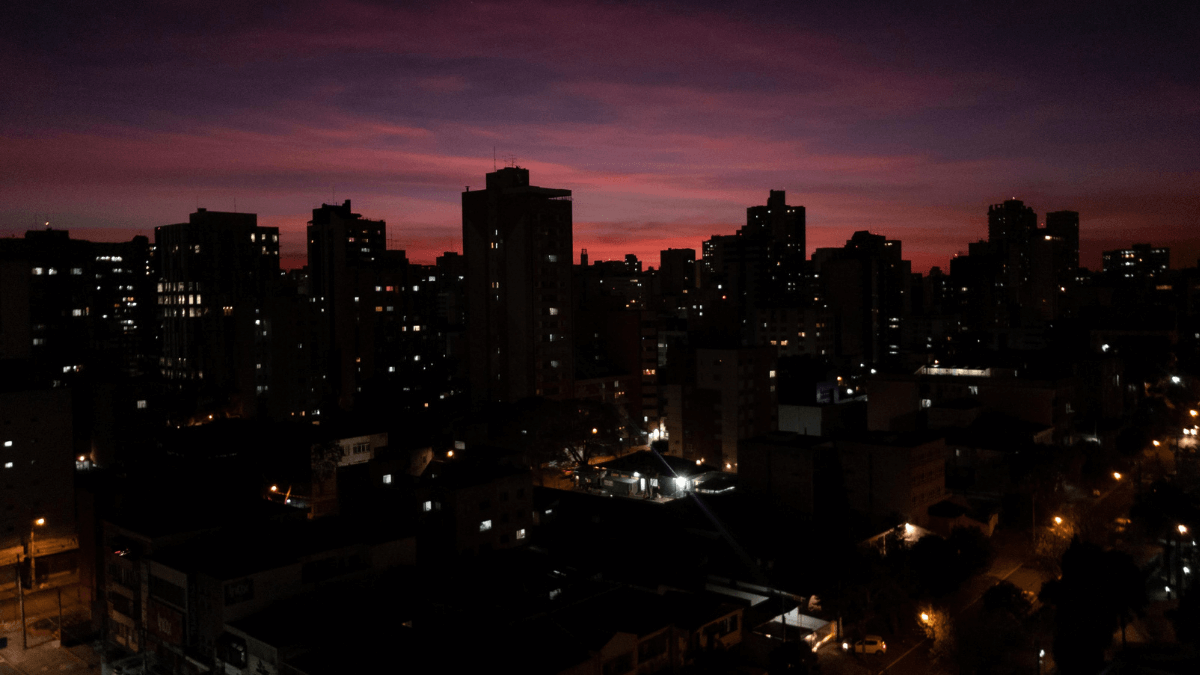 Céu noturno de Curitiba pintado em tons de rosa e roxo após o pôr do sol, com silhuetas de edifícios urbanos e luzes começando a brilhar.