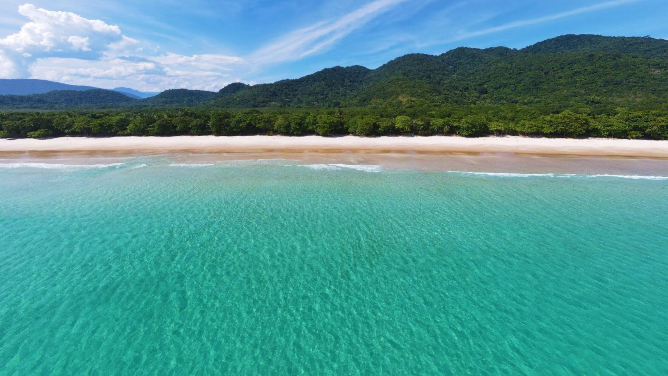 Vista aérea de uma extensa praia de areia clara com águas azul-turquesa que se estendem até a linha do horizonte, separada por uma densa vegetação tropical. Céu claro com nuvens dispersas complementa a serenidade da cena.