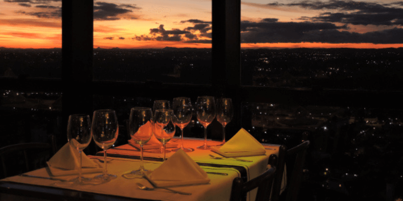 Mesa de restaurante com taças vazias tem vista para um pôr do sol laranja brilhante sobre Curitiba, oferecendo uma experiência de jantar com vista panorâmica.