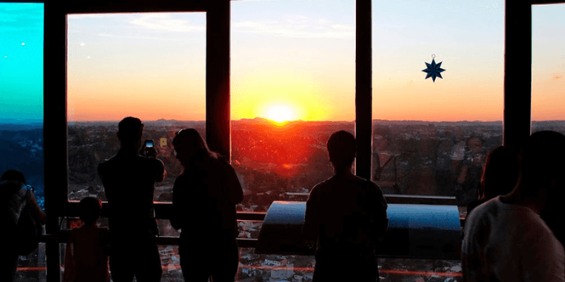 Visitantes observam o sol se pôr no horizonte de Curitiba, capturando o momento através das grandes janelas de um observatório.