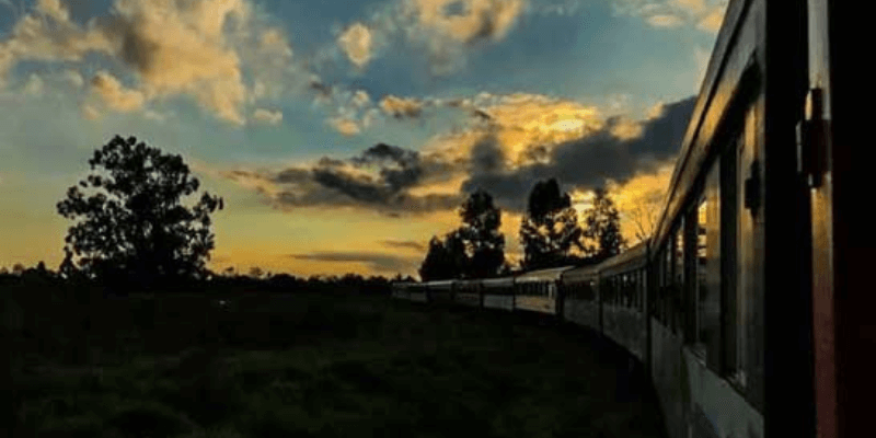 Trem viajando ao entardecer em Curitiba com o céu colorido de amarelo e azul, destacando as nuvens e o contorno das árvores ao longo da ferrovia.