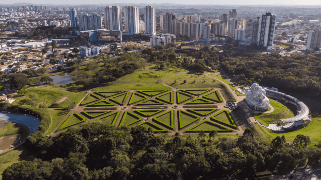 Fotografia aérea mostrando um parque em primeiro plano com padrões simétricos de jardins bem cuidados, uma grande estrutura de vidro que parece uma estufa, e um prédio moderno com um design arquitetônico distinto que se assemelha a um olho. Ao fundo, o skyline urbano de Curitiba com edifícios altos.