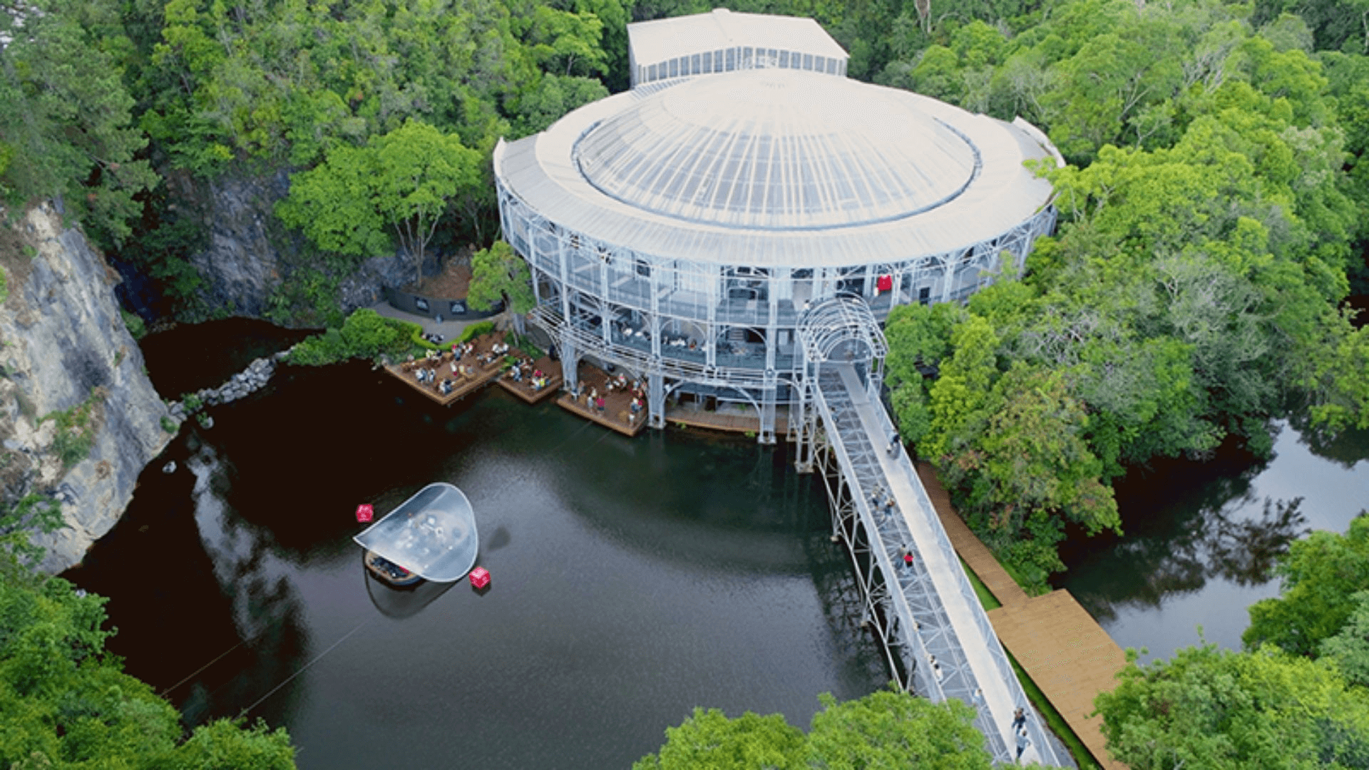 Imagem aérea da Ópera de Arame, mostrando sua estrutura metálica circular única que se sobressai sobre um lago. A natureza circundante é densa e exuberante, com árvores verdes que realçam a construção e a paisagem natural ao redor.
