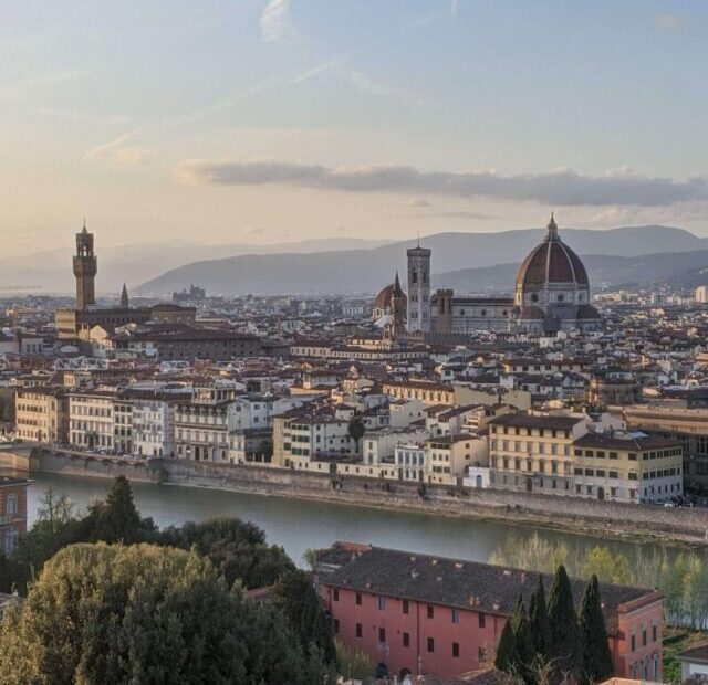 Paisagem panorâmica de Florença ao entardecer, mostrando uma vista aérea da cidade com seus prédios históricos e o rio Arno. O céu tem tonalidades suaves de azul e laranja.