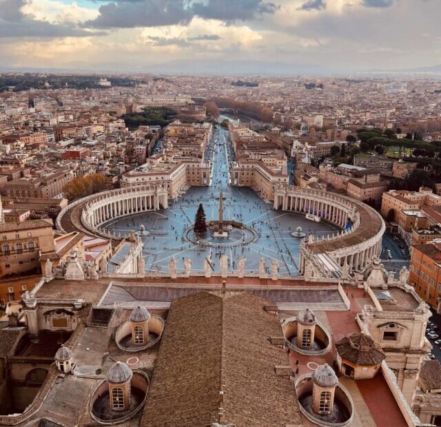 A imagem oferece uma vista aérea espetacular da Praça de São Pedro no Vaticano, mostrando a simetria radial das colunatas que abraçam o espaço oval central com a obelisco egípcio ao centro. Ao fundo, a cidade de Roma se estende até o horizonte sob um céu parcialmente nublado ao entardecer.