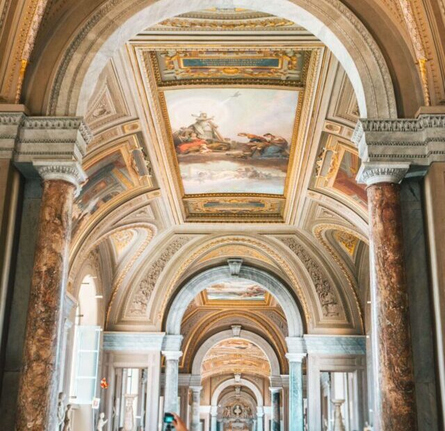 imagem mostra um corredor abarrotado de visitantes no Museu do Vaticano, ladeado por colunas e estátuas clássicas. Os tetos são ricamente decorados com afrescos e molduras douradas, exibindo cenas históricas e figuras mitológicas.