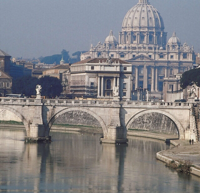 Uma paisagem clássica de Roma, mostrando o Rio Tibre com a Ponte Sant'Angelo em primeiro plano e a imponente Basílica de São Pedro ao fundo. A fotografia captura a serenidade do rio e a majestade arquitetônica da cidade.